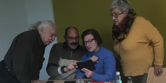 Vier Menschen sitzen am Frühstückstisch und betrachten ein Smartphone
