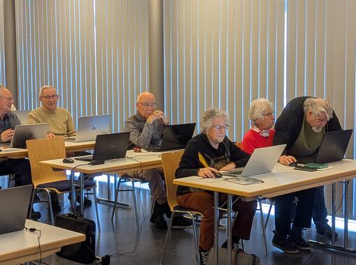 Eine Gruppe von lernenden Seniorinnen und Senioren sitzt an Tischen. Alle nutzen jeweils einen Laptop. Am vorderen Tisch erklärt ein Mann zwei Frauen etwas.