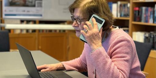 Frau C. sitzt am Laptop und telefoniert gleichzeitig mit dem Handy. Dahinter eine Bücherwand.