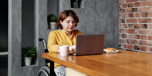 Eine Frau sitzt am Tisch in ihrem Rollstuhl. Davor steht ein aufgeklappter Laptop, auf den sie tippt. 