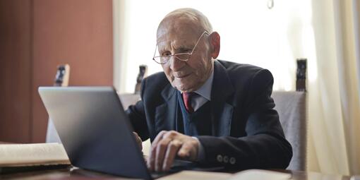 Ein älterer Mann sitzt am Tisch und tippt auf einen Laptop. Neben ihm liegen Bücher.