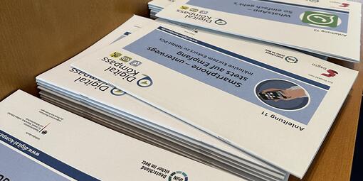 Anleitungen des Digital-Kompass liegen auf einem Regal. Darunter Online-Banking, Exkurs Smartphone- und Tablet sowie WhatsApp.