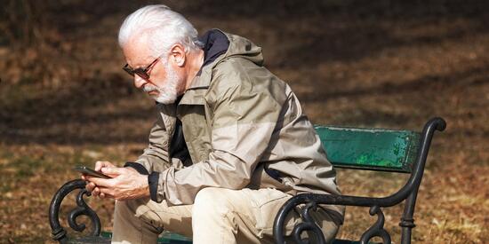 Ein älterer Herr sitzt mit seinem Smartphone in der Hand auf einer Parkband.