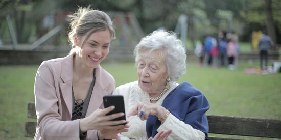 Eine jüngere und eine ältere Frau sitzen gemeinsam auf einer Bank im Park. Sie schauen fröhlich gemeinsam auf ein Smartphone. Die ältere Dame zeigt auf das Smartphone und sagt etwas dazu.