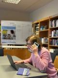 Frau C. sitzt am Laptop und telefoniert gleichzeitig mit dem Handy. Dahinter eine Bücherwand.