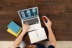 Ein Laptop steht auf einem braunen Holz-Tisch, links daneben liegen Notizbücher. Eine Person sitzt daran und hält eine Teetasse in der Hand. Das Bild wurde von oben fotografiert.