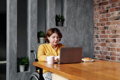 Eine Frau sitzt am Tisch in ihrem Rollstuhl. Davor steht ein aufgeklappter Laptop, auf den sie tippt. 