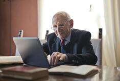 Ein älterer Mann sitzt am Tisch und tippt auf einen Laptop. Neben ihm liegen Bücher.